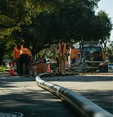 Workers in orange safety vests perform maintenance on a residential street with equipment and traffic cones, a large hose or pipe extends across the road.