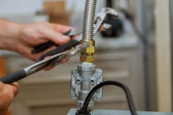 A person uses pliers to tighten a flexible metal gas line connection to a valve, with tools and appliance parts visible.