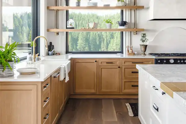 Modern kitchen with light wood cabinets, white countertops, open shelves with plants, a large window with a forest view, and a farmhouse sink.