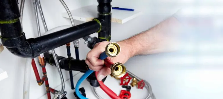 A person's hand holds two water hoses with brass fittings, connected to plumbing under a sink.