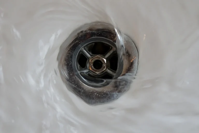 Close-up of water swirling around a metal drain in a sink, creating a small vortex as it flows downward.