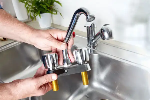 Person holding a detached kitchen faucet with two handles and brass threads above a stainless steel sink, with another faucet and a plant in the background.